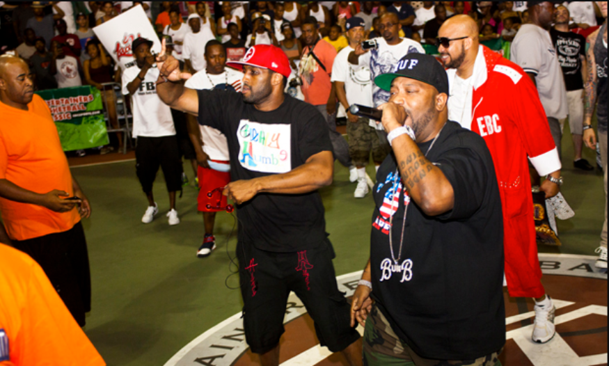 A lively basketball event with a packed crowd, featuring hip-hop artists performing on the court. One performer is holding a microphone while another gestures to the audience, creating an energetic atmosphere.
